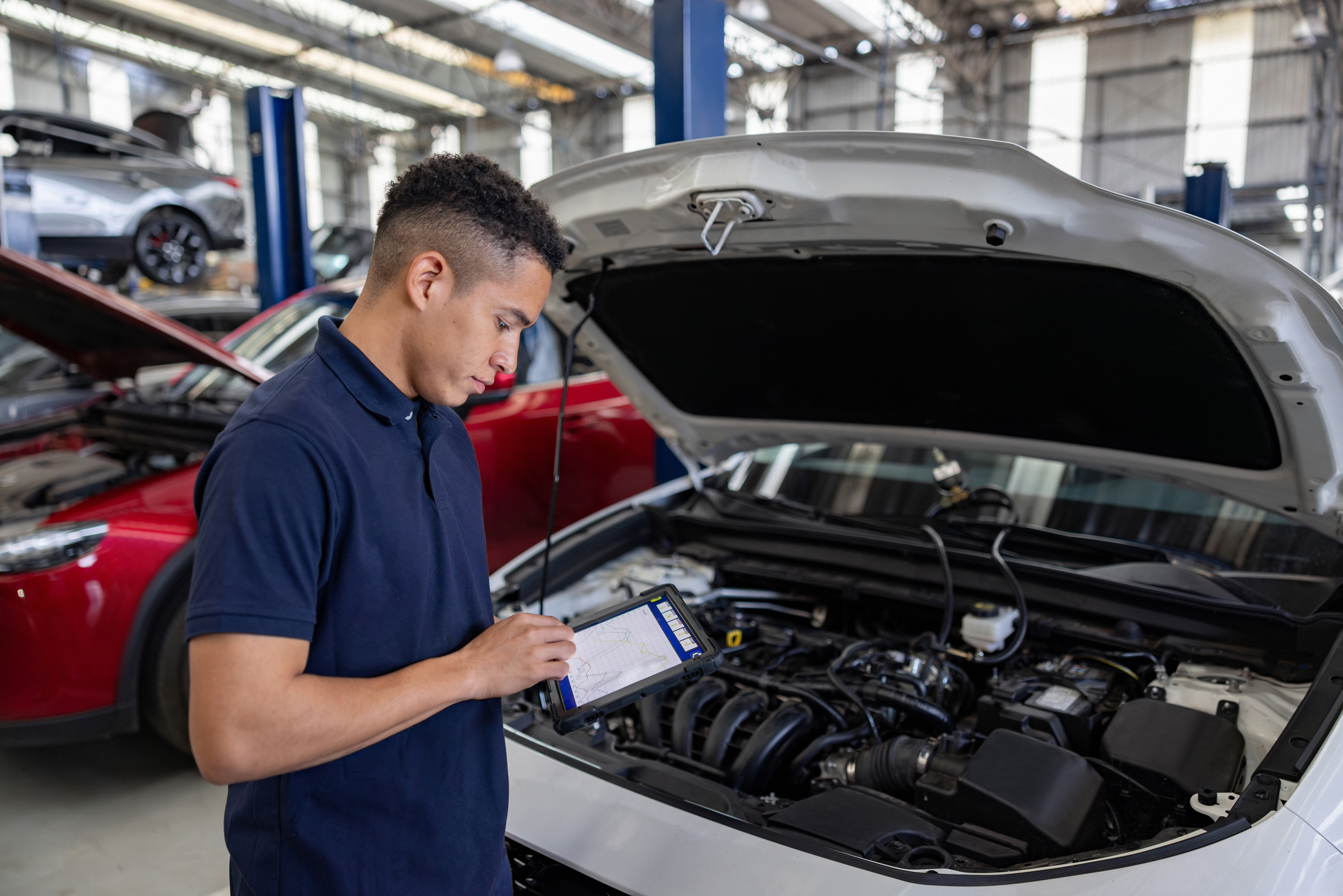Man standing in a garage, holding a tablet computer and looking at the engine of a car