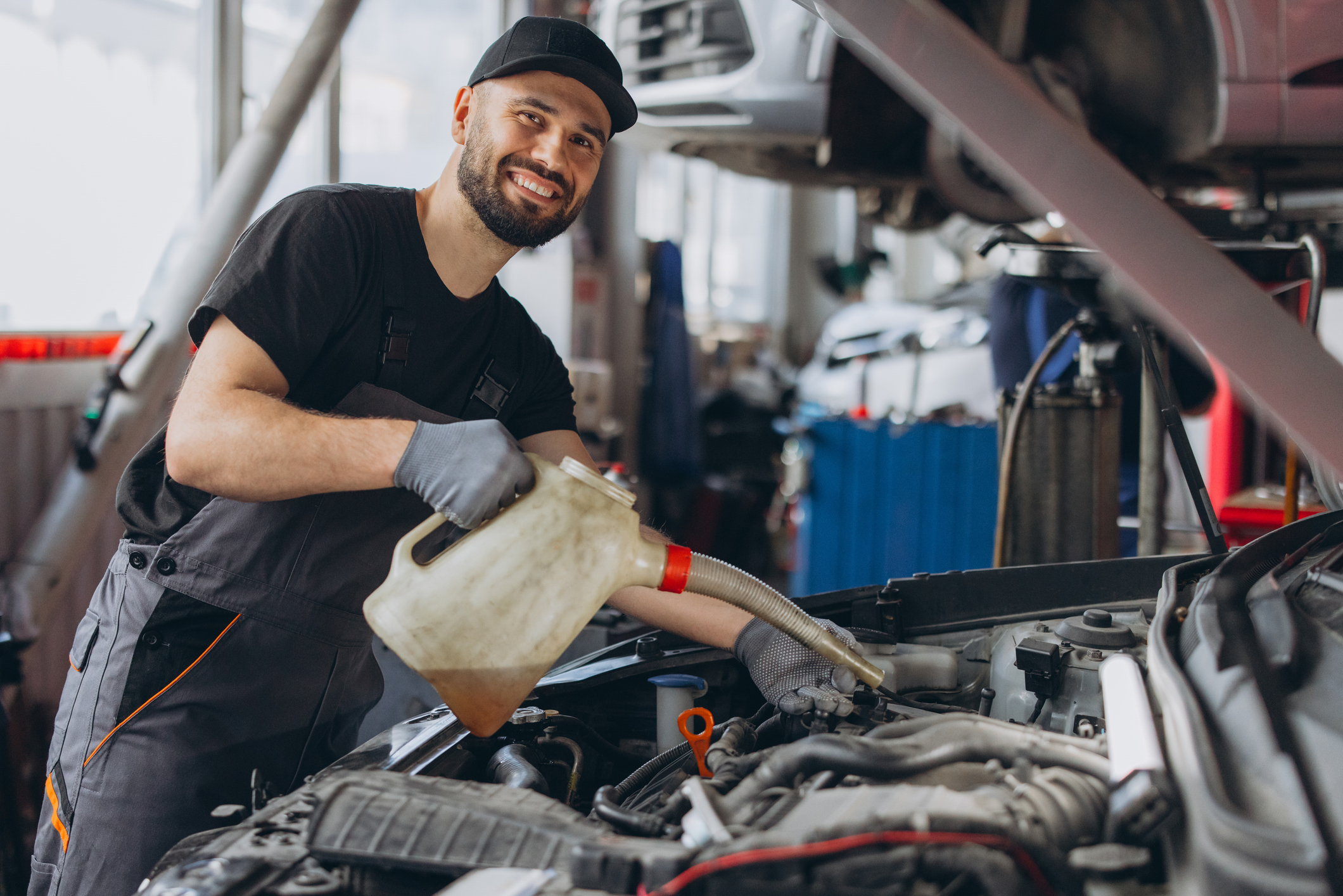 Mechanic wearing a cap and gloves, holding a can of oil and pouring it into the engine of a car
