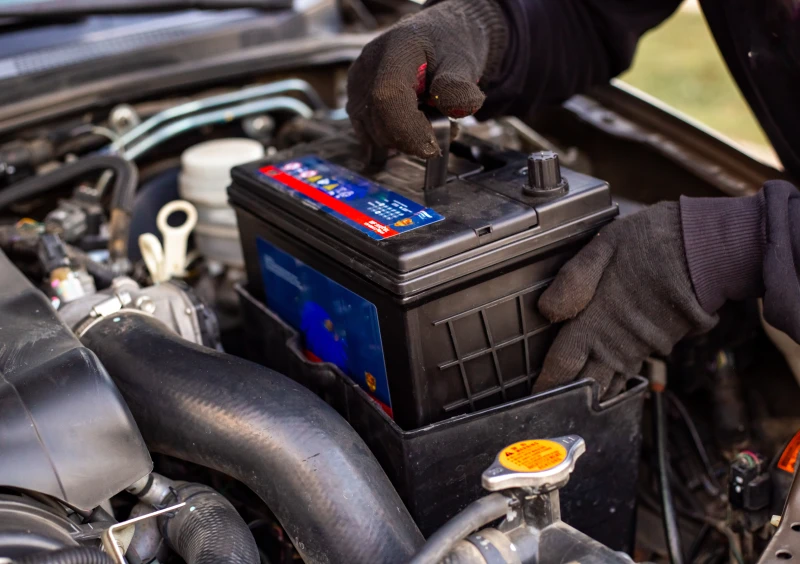 Person wearing gloves and holding a car battery in front of a car, with the background blurred